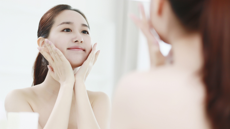 Young Woman Looking In Mirror, Touching Face